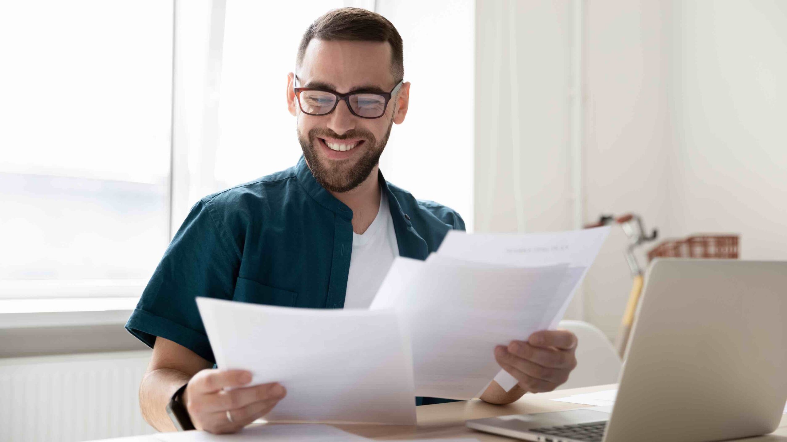 A nice businessman holding and reading documents, with good news near laptop.