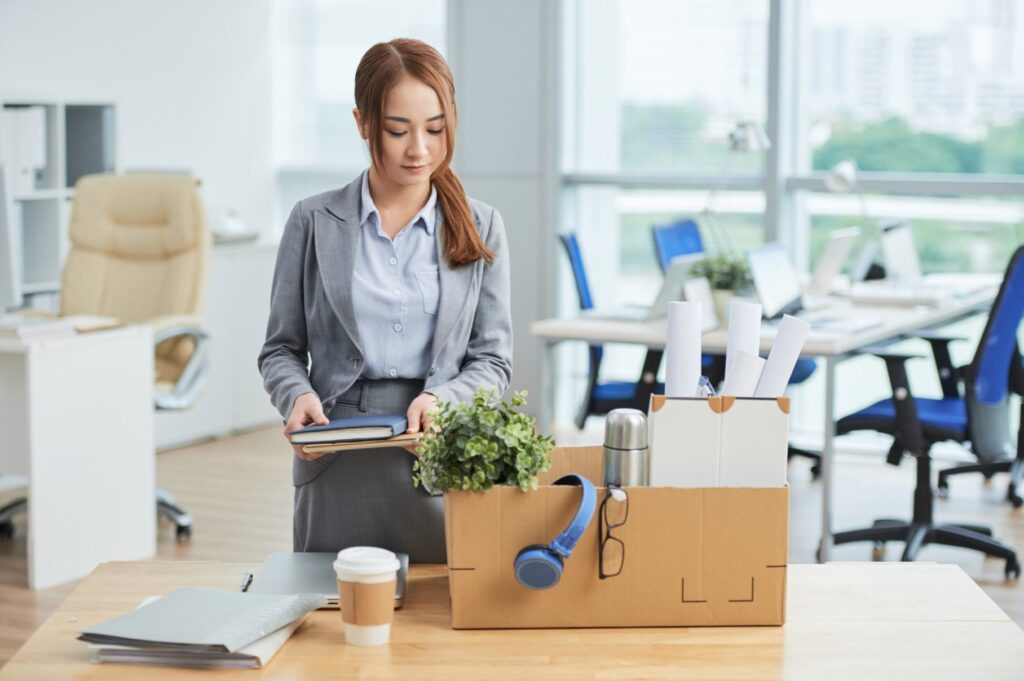 Asian woman standing at deks in office with belongings in cardboard box