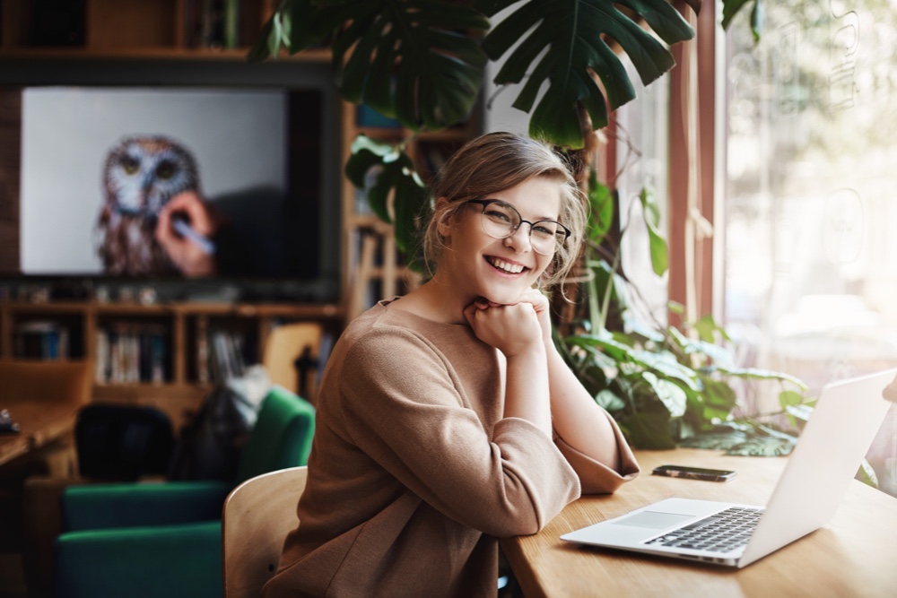 Charming and carefree female in glasses, sitting near window and laptop, leaning on hands