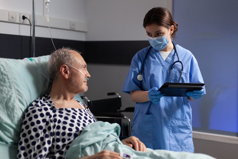 Medical assistant with protection mask checking treatment of senior man reading notes on tablet