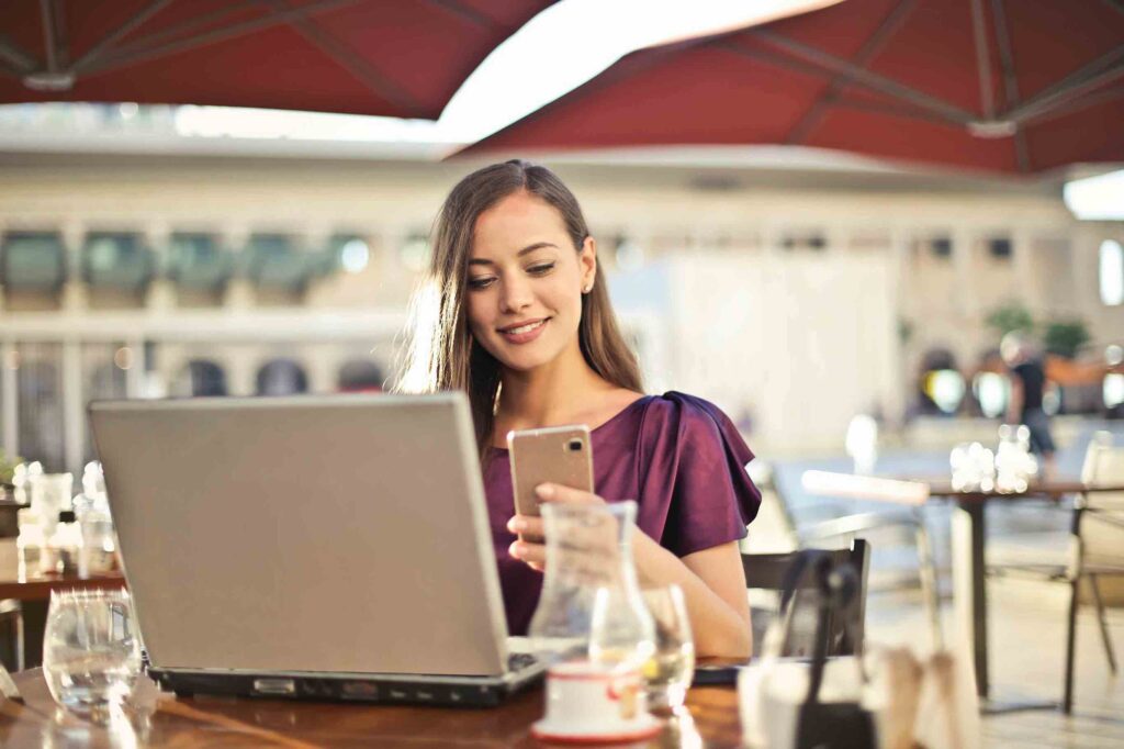 Woman in restaurant in front of laptop checking resume samples on her phone