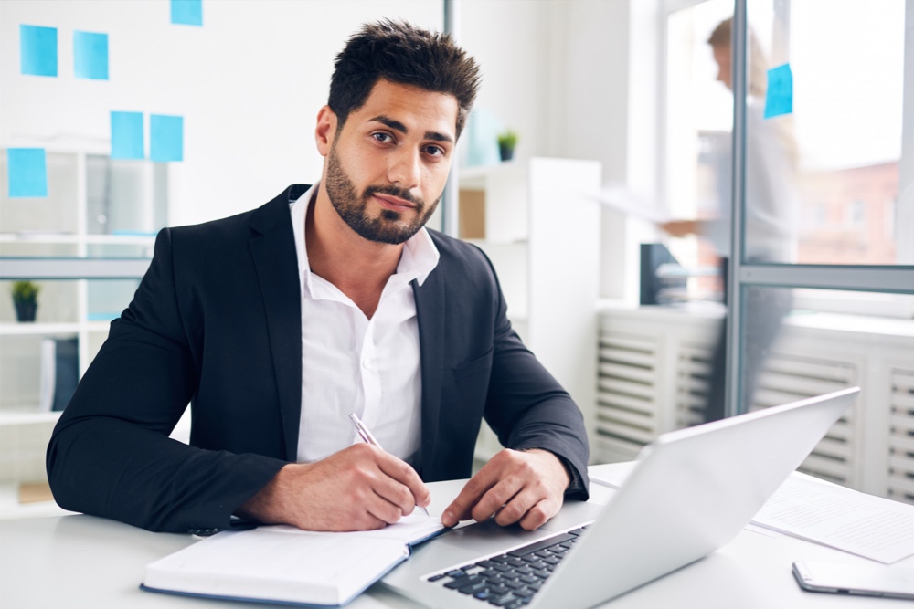 Young accountant in front of laptop putting something down in a notebook