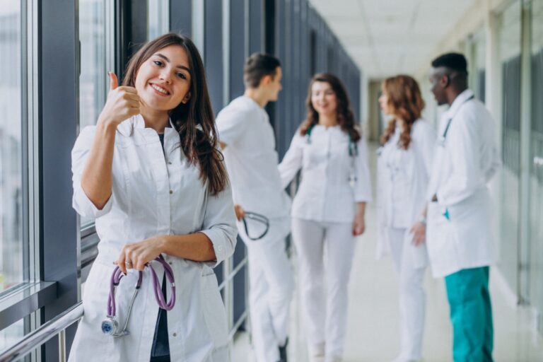 Young female medical assistant showing thumbs up standing in hospital corridor