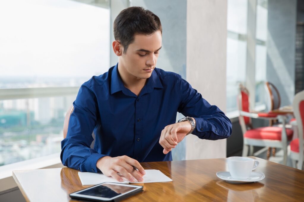 Young, serious employee looking at his watch while writing something in a notebook