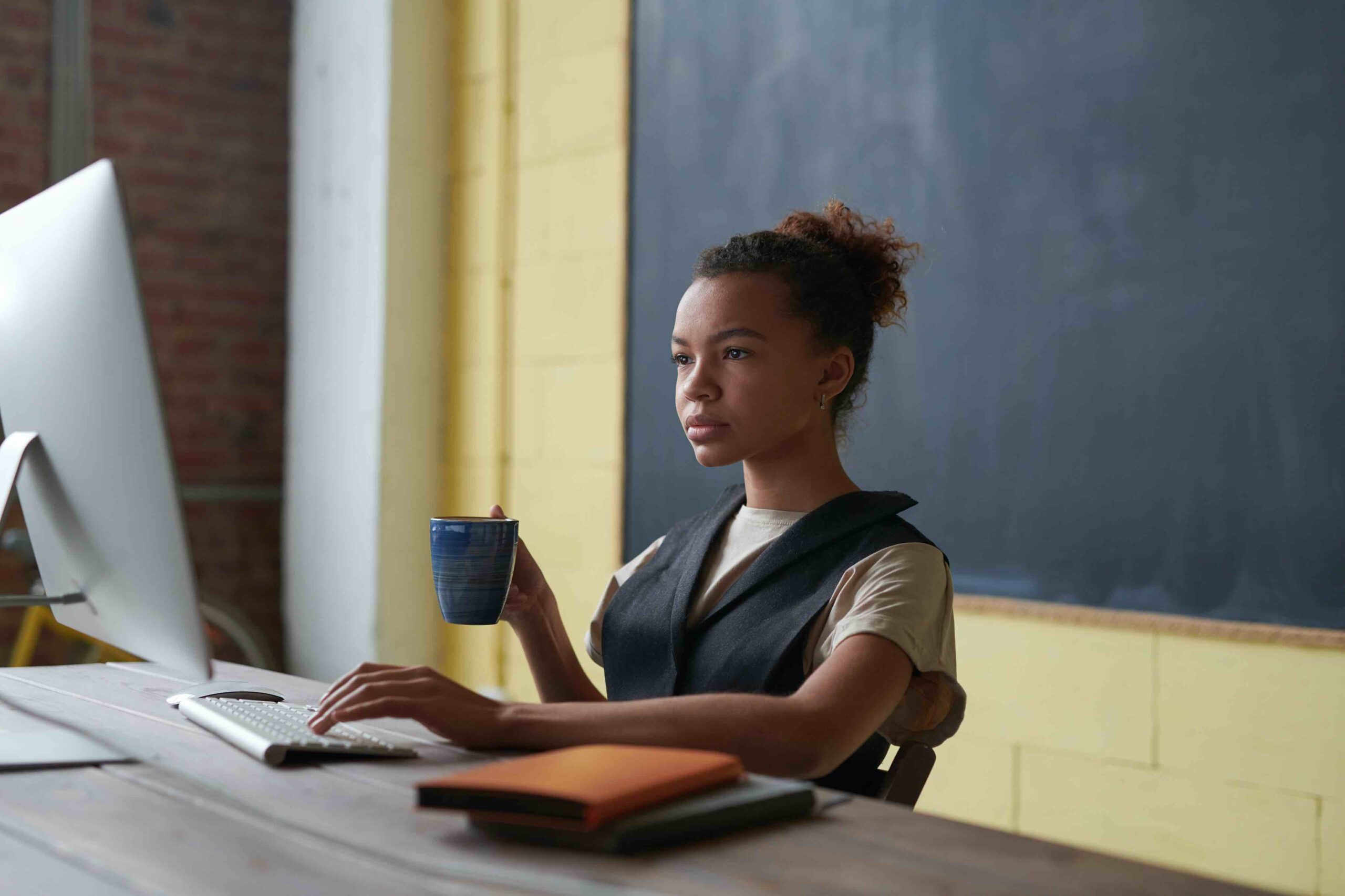 Young teacher at her desk looking at computer while holding cup