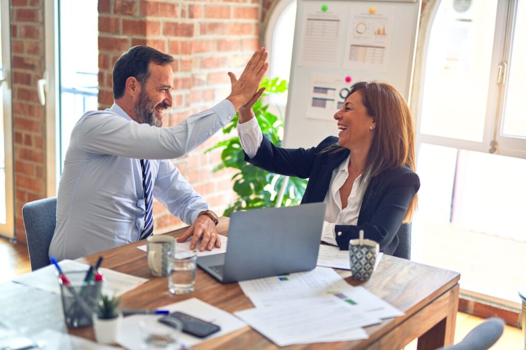 Male and female colleague in business clothing sitting at a desk, laughing and giving each other a high five