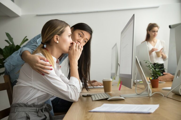 woman sitting in front of a pc screen, looking slightly desperate, being comforted by another woman who is holding her shoulders with her hands