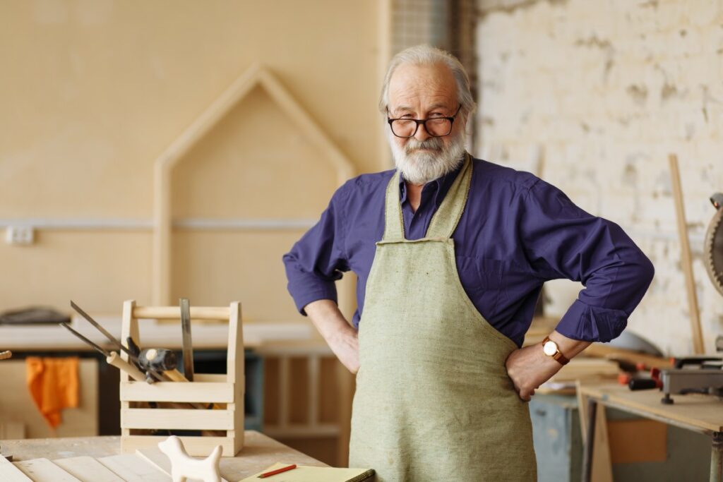 elderly man with glasses wearing a purple button down and and a beige apron, stemming his hands into his hips