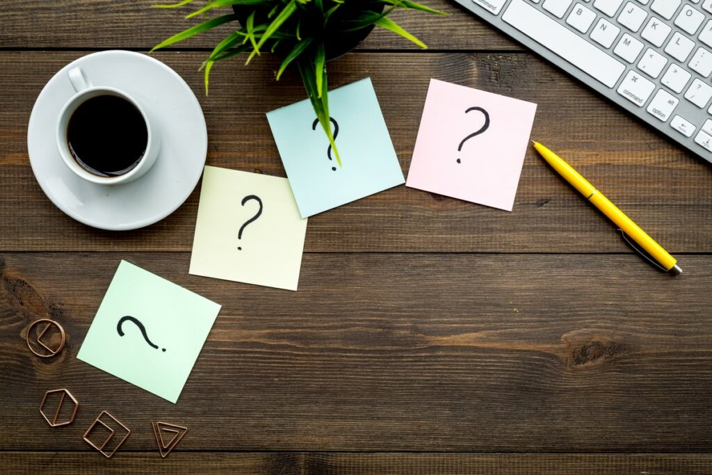 top view of a dark wooden table with a cup of coffee and 4 post it's in different pastel shades with black questionmarks on them