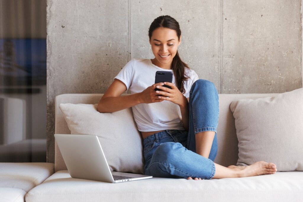Woman sitting on a white couch with a laptop on the couch and a smartphone in her hand smiling at the screen