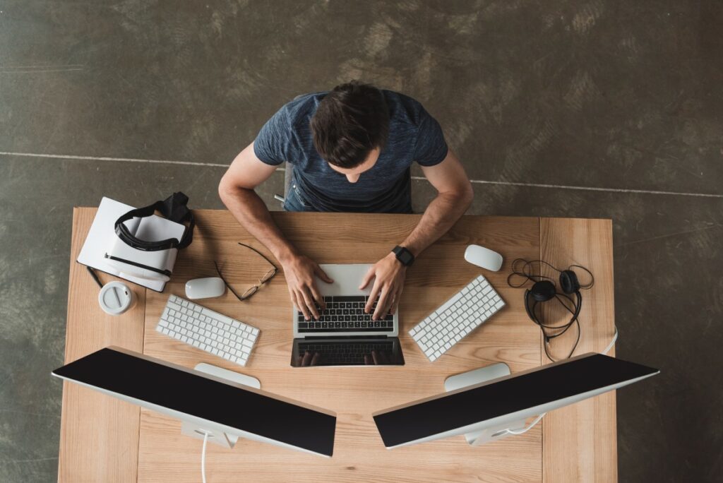 Top view of a man working in a digital workplace on laptop, two screens, two keyboards, and more