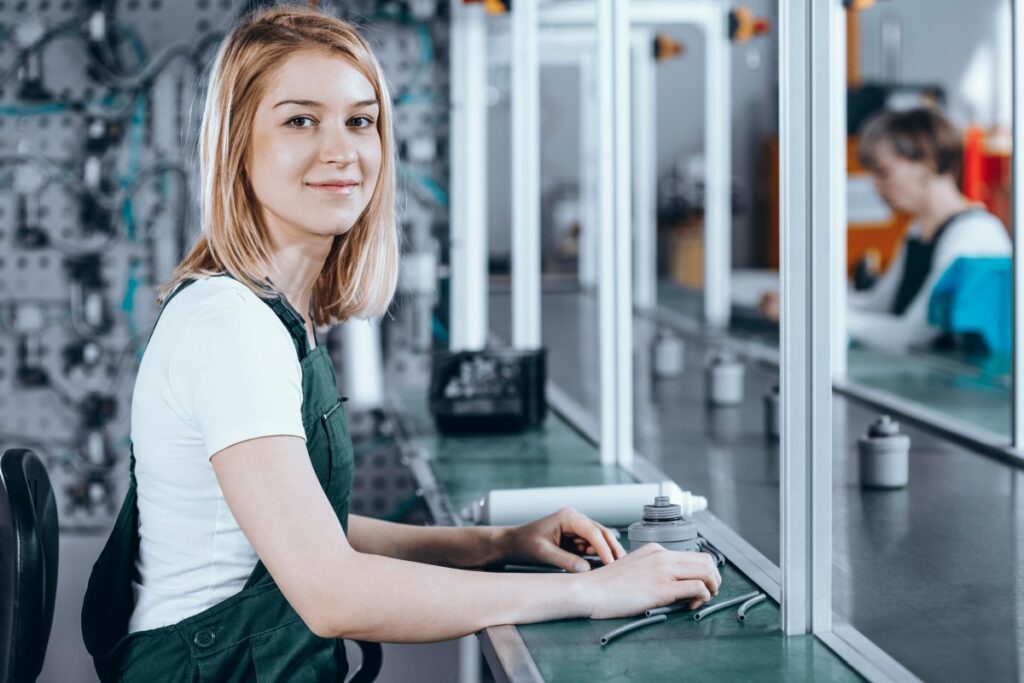 woman standing in a workshop with white shirt and green overalls, smiling 