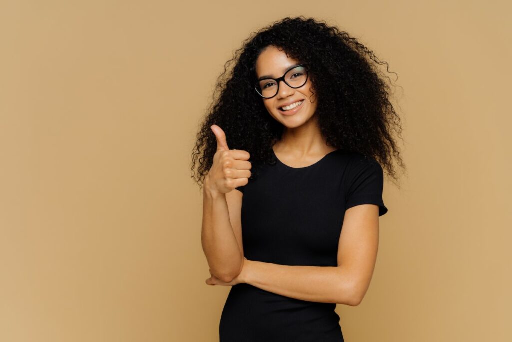 Woman dressed in black with glasses smiling and showing a thumbs up in front of a beige background