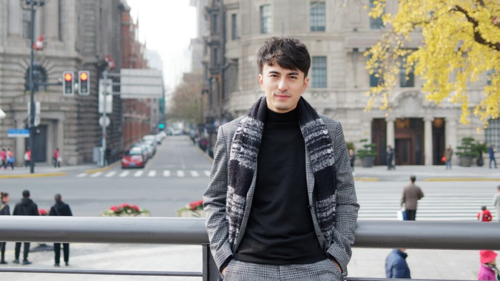 Man with dark hair standing on the street in winter, wearing a black turtleneck with grey suit and a grey-black scarf