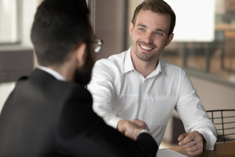 Two men shaking hands sitting at a table, wearing a white button-down, confidence appearance, smiling confidently while mastering his body language during a job interview