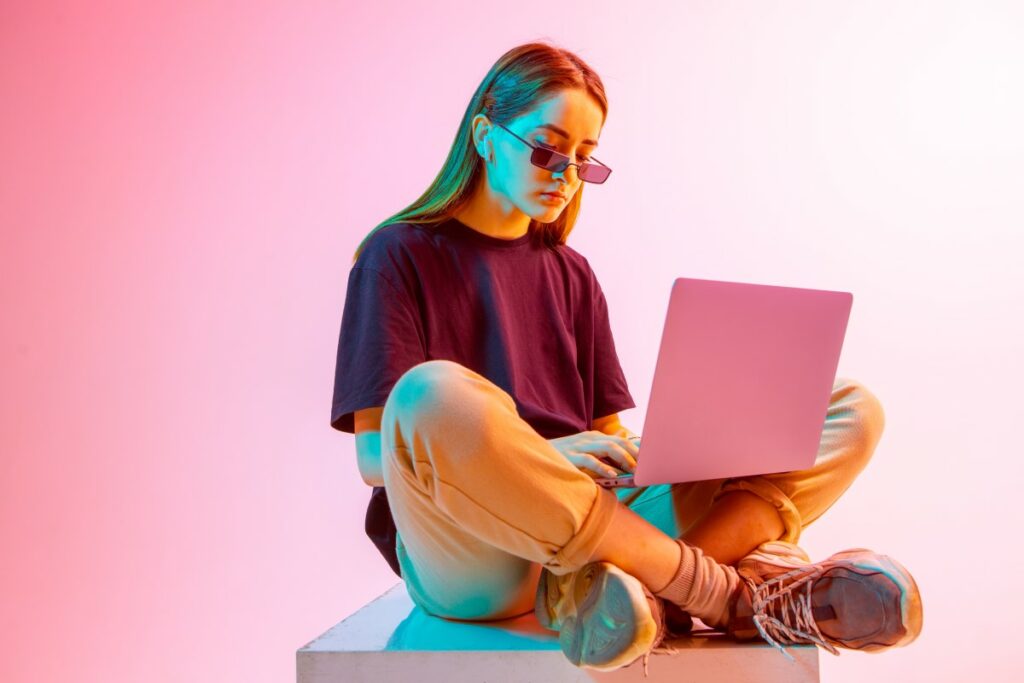 girl with purple shirt and purple sunglasses sitting on the floor, legs crossed, with a pink laptop on her knees