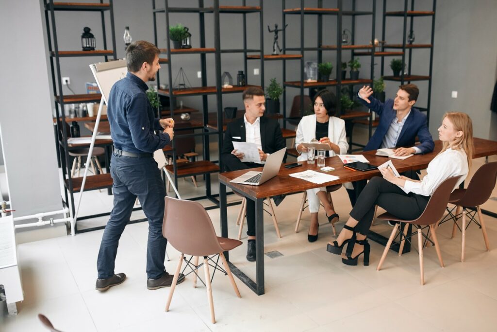 five people in a modern cafe like office space, 4 sitting at a table, one standing in front of them on a flipchart