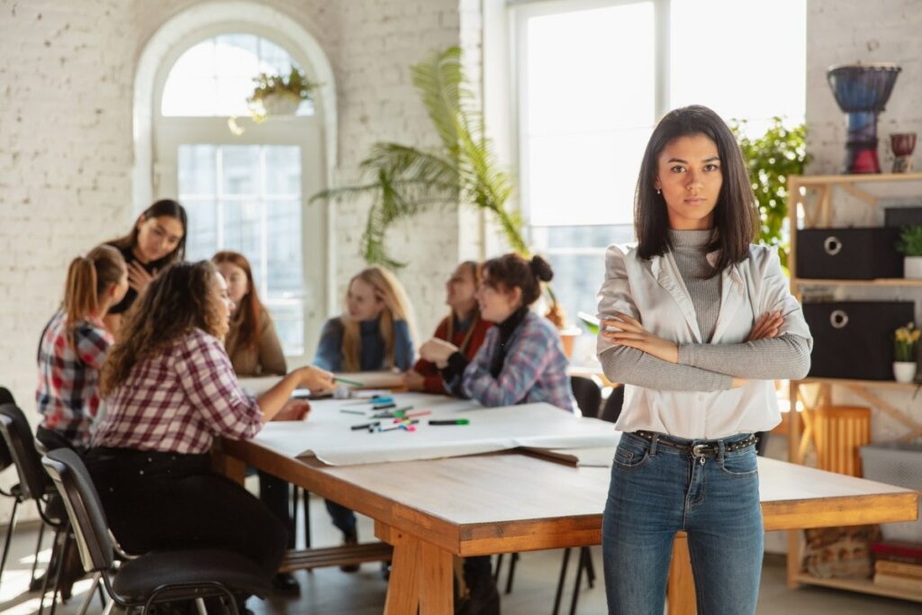 a woman stands in the foreground and looks stern, while in the background a group of women learn in a circle