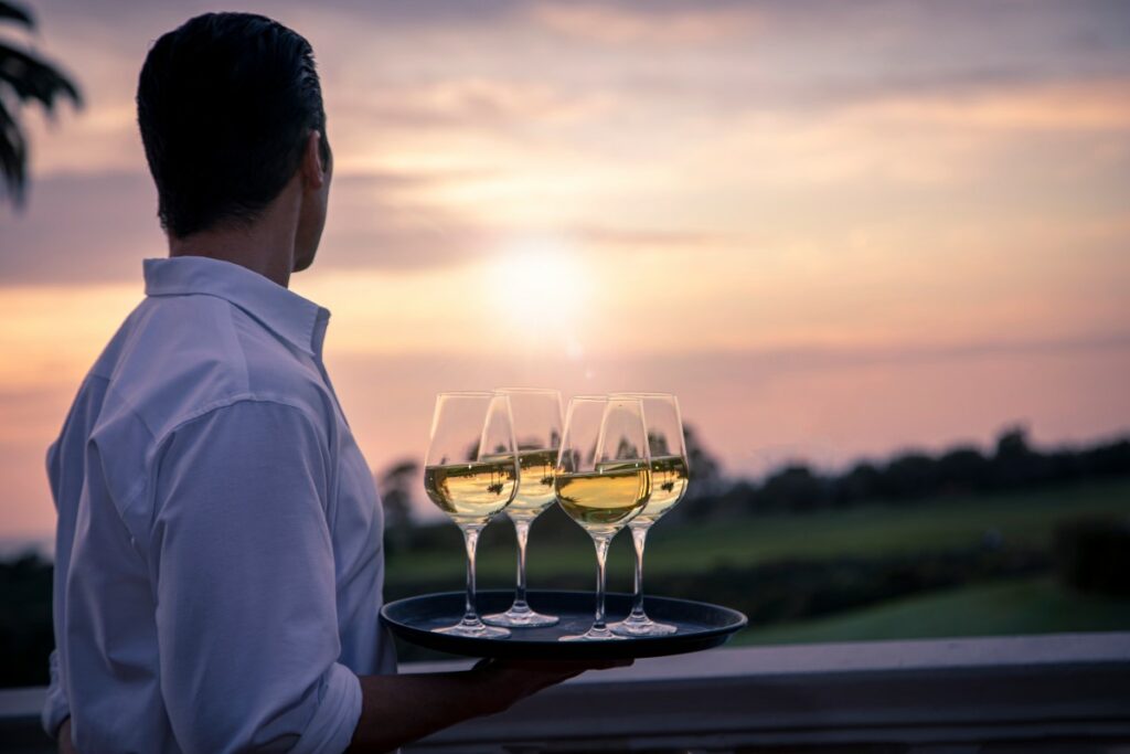 man in a white button down holding a trey with wine glasses filled with white wine looking into the sunset