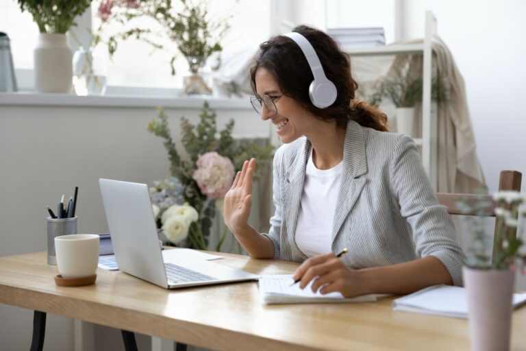 woman with a light grey blazer wearing glasses and white headphones sitting on a desk waving in front of her laptop