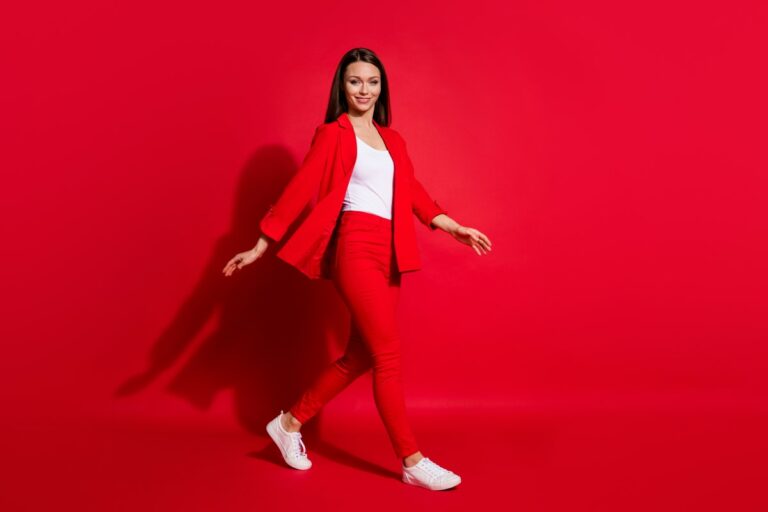 woman dressed in a red pantsuit, white shirt and white sneakers walking in front of a red background