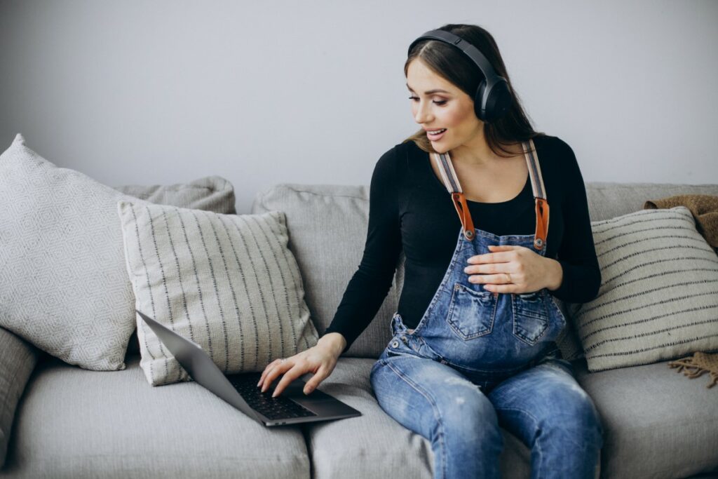 pregnant woman sitting on the couch wearing headphones and working on a laptop