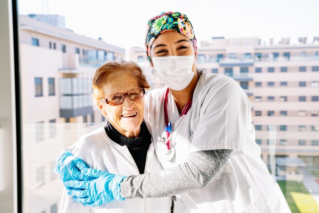 nurse with mask and blue gloves hugging an elderly woman, both are smiling