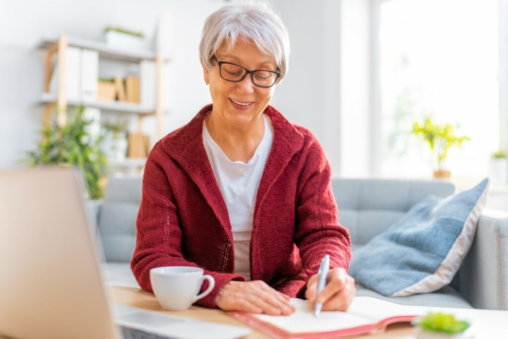 elderly woman with glasses wearing a white shirt and a red jacket writing something down, smiling