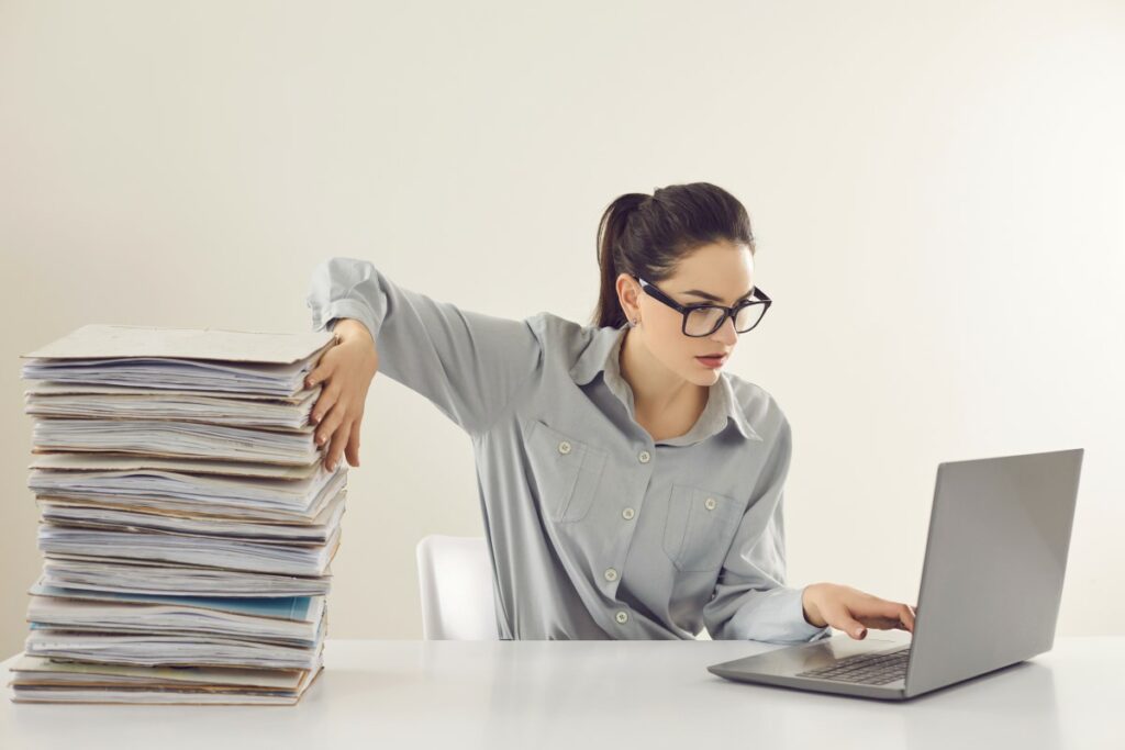 A woman pushes files aside with her right hand while she uses her laptop with her left hand in concentration.