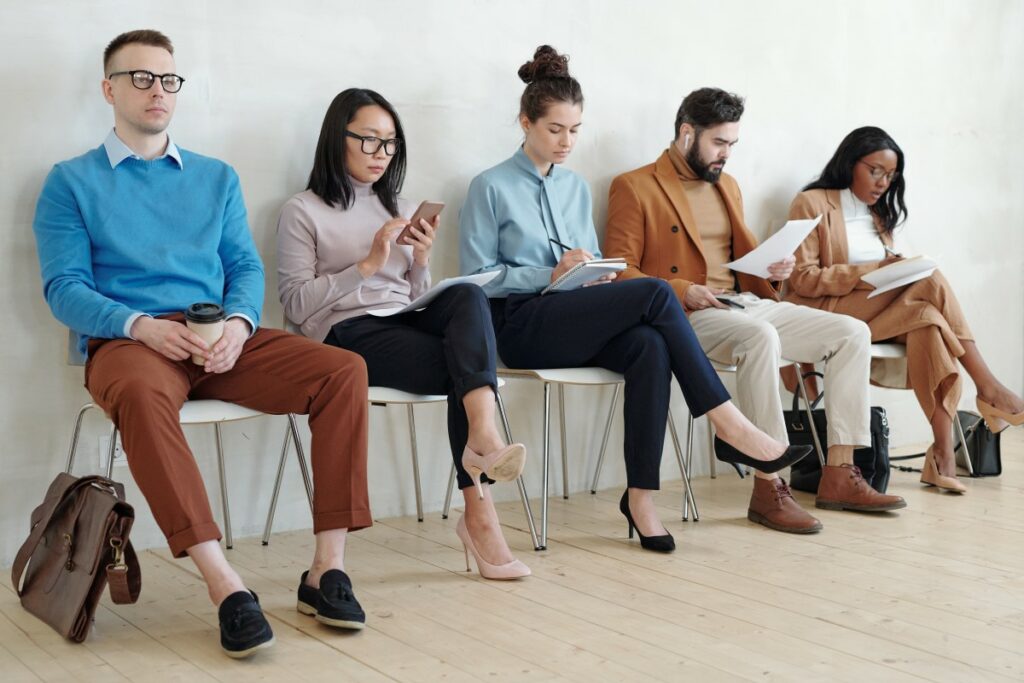 five people, men and women, sitting on chairs, reading on phones or in books, writing something down, drinking coffee, waiting for their job interviews