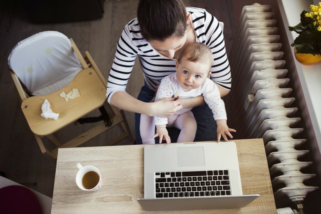 top view of woman sitting at a desk, a laptop and a cup of tea on the desk, a baby on her lap