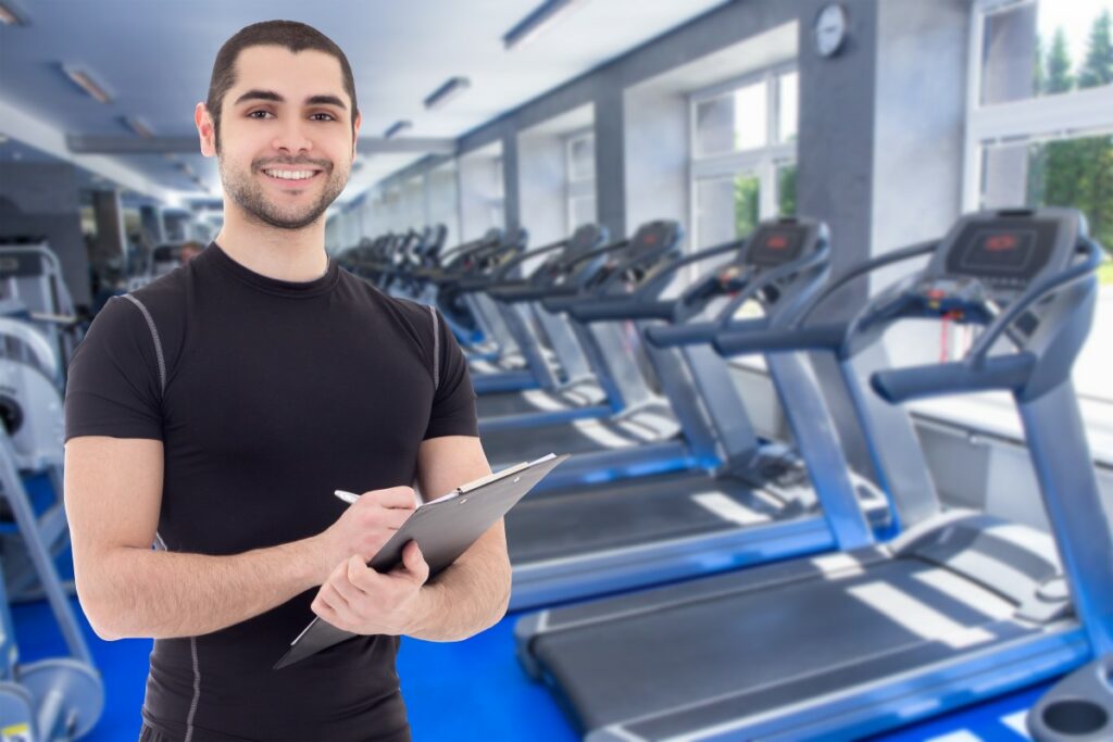 man in a tight black shirt standing in a gym in front of a bunch of treadmills, holding a clipboard and pen, smiling