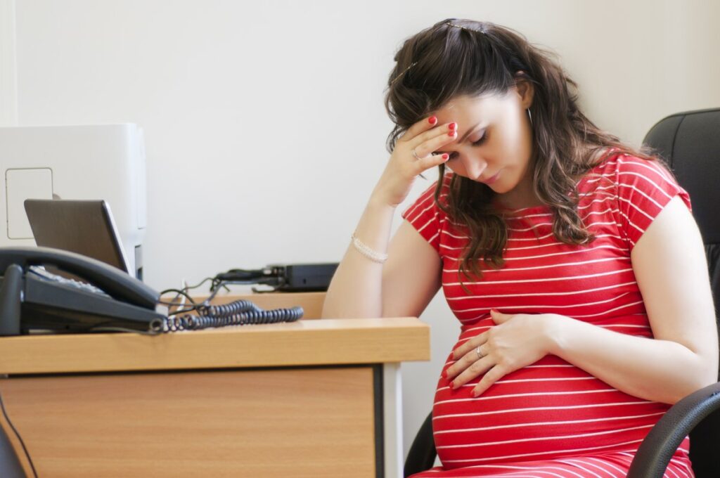 pregnant woman wearing a red shirt sitting at a desk at the office, holding her head and her belly, looking stressed and sad