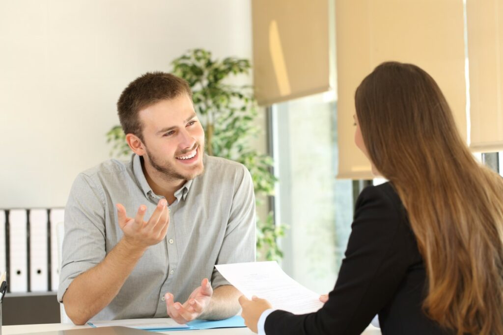 Two people sitting at the table during a job interview, one holding a resume, the other one talking confidently and showing confidence with his body language