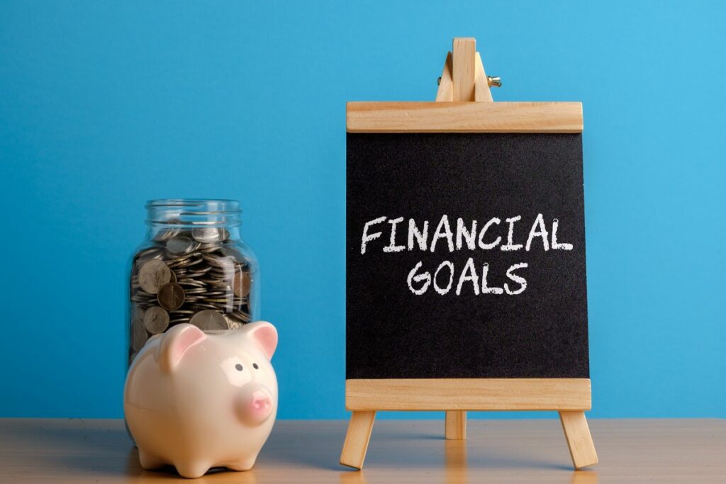 tiny blackboard with financial goals written on it with white chalk, piggy bank and jar with coins standing next to it