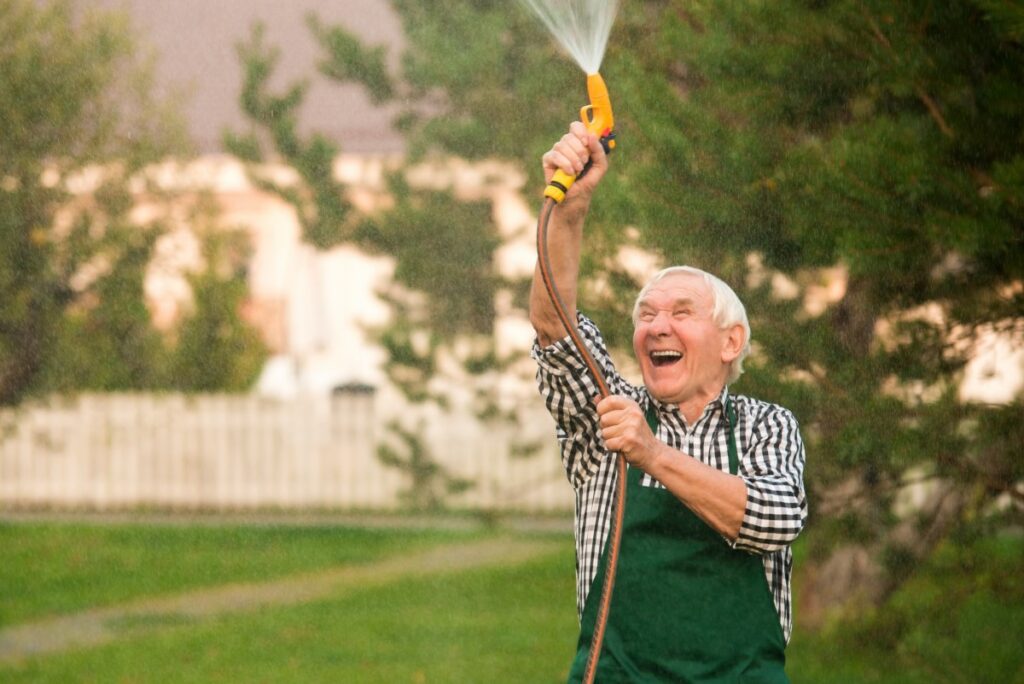 elderly man wearing a plaid shirt and green overalls spraying water up in the air with a garden hose, laughing