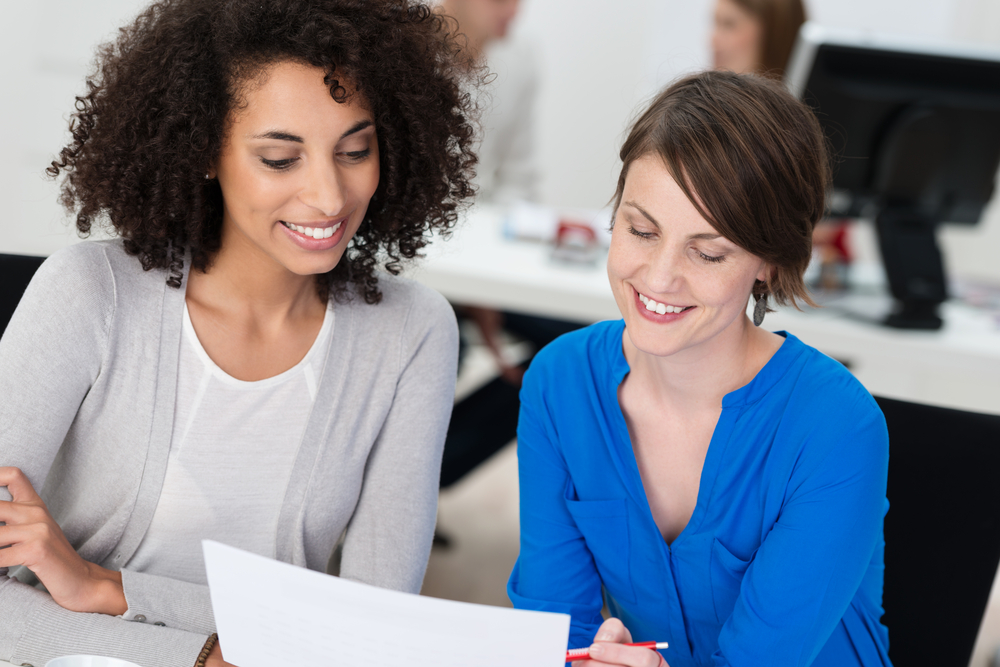 Two,Smiling,Businesswomen,Working,On,A,Document,As,They,Sit