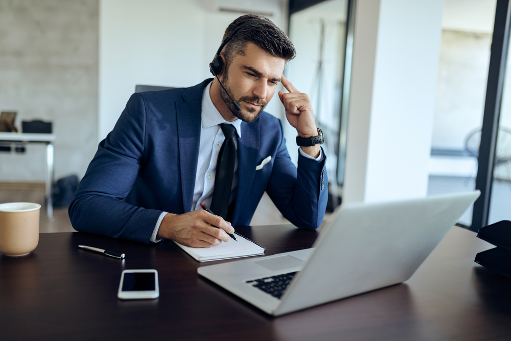 A young business man is taking notes during an online meeting.